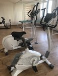 Wnętrze dziennego domu pomocy. Pomieszczenie rehabilitacji. Na pierwszym planie dwa rowery stacjonarne. W głębi klatka rehabilitacyjna ze stołem rehabilitacyjnym